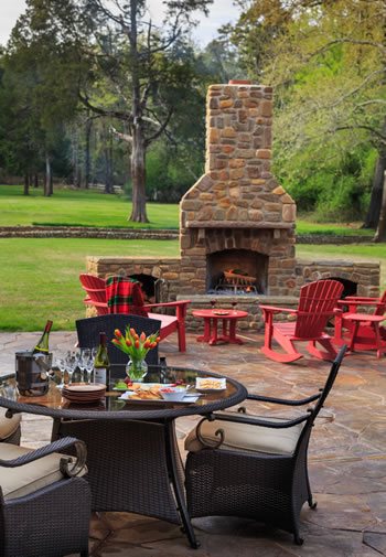 Outdoor patio with table and chairs in the foreground and a stone fireplace in the background with green grass.